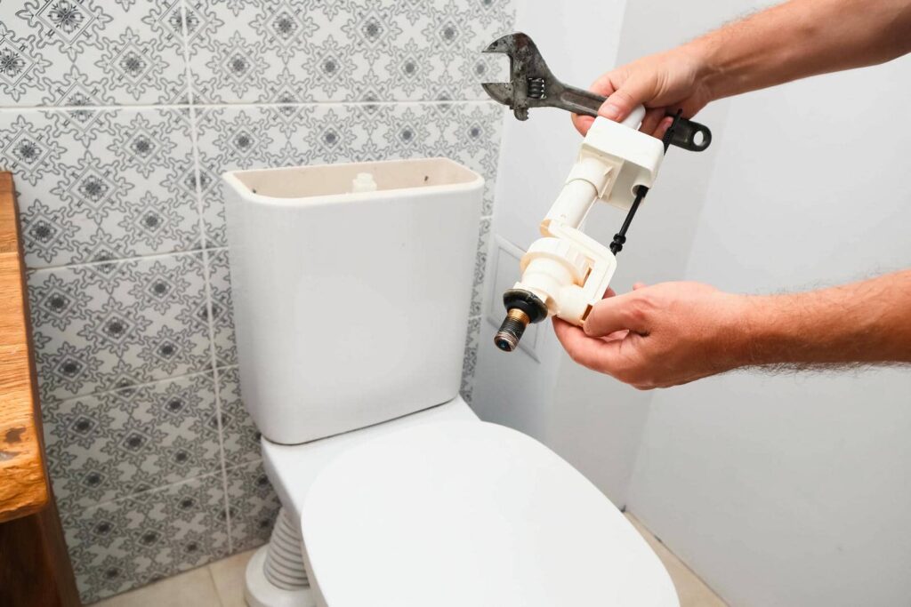 Hands holding a toilet fill valve and an adjustable wrench next to an open toilet tank.